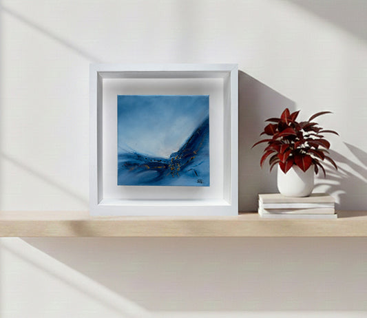 Original blue abstract art by Sinead Guilfoyle, Emergence 1, framed in white on a minimalist shelf. Contemporary spiritual wall art for home decor featuring textured gold leaf and oceanic blue tones.