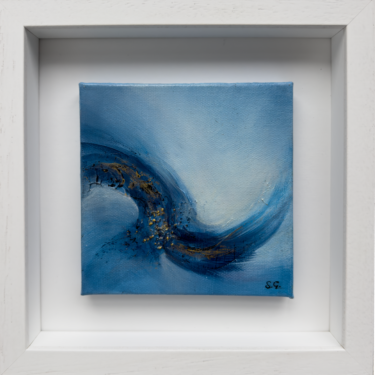 A square abstract painting, Emergence 2 by Sinéad Guilfoyle, with sweeping blue brushstrokes and gold leaf accents, titled Emergence 1, showcased in a white wooden shadow box frame.